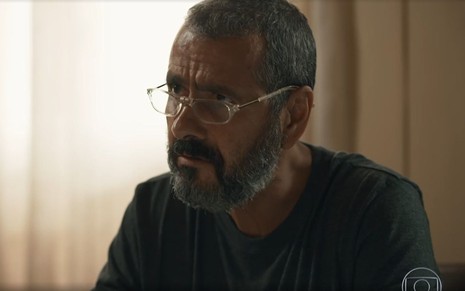 Em cena de Renascer, Marcos Palmeira está com a expressão séria; ele usa blusa escura e óculos de grau
