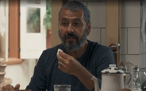 Em cena de Renascer, Marcos Palmeira está falando com alguém, enquanto tem algo de comer na sua mão