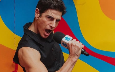 Reynaldo Gianecchini faz pose com microfone em cenário com fundo colorido