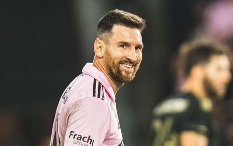 Lionel Messi usa camisa rosa do Inter Miami; ele olha para o lado e sorri durante jogo