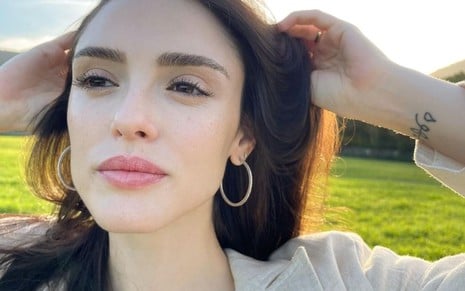 Isabelle Drummond usa um blazer branco e está com as mãos nos cabelos; ela caminha sobre um campo verde