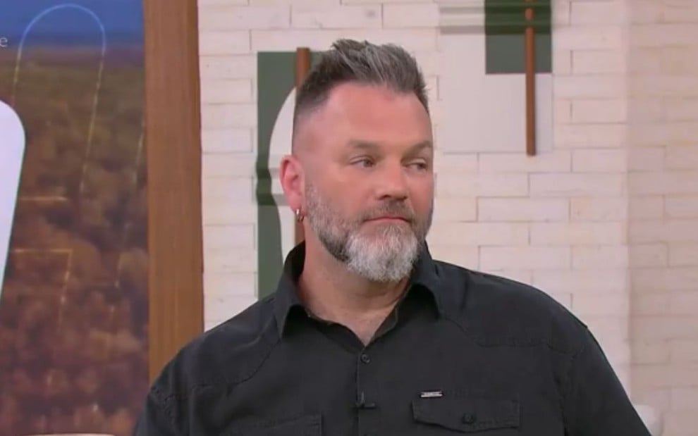 A imagem mostra um homem de barba grisalha e cabelo curto, usando uma camisa preta. Ele está sentado em um estúdio de TV, com objetos de mesa à frente.