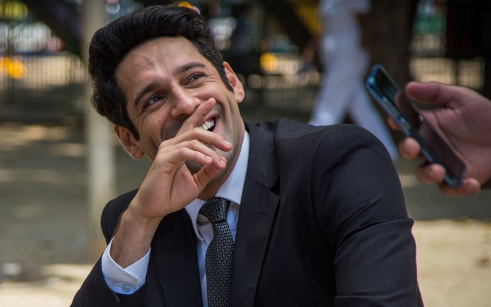 O ator João Baldasserini sorrindo, com a mão na frente da boca, de terno e gravata