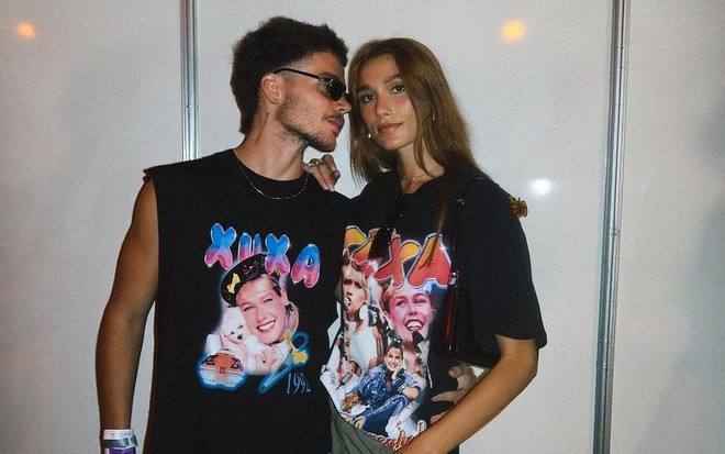 De óculos escuros, João Lucas olha para Sasha Meneghel, que faz carão para a câmera; os dois usam camisetas com estampa de Xuxa