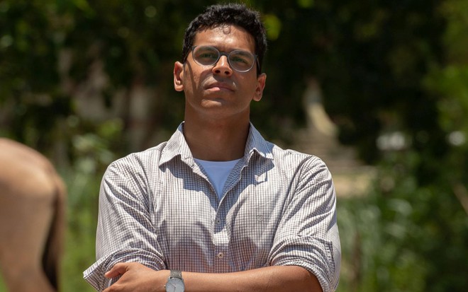 Caracterizado como seu personagem em Renascer, Renan Monteiro usa blusa quadriculada de botão e uma camisa branca por baixo; ele usa óculos