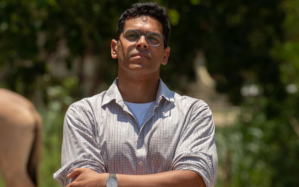 Caracterizado como seu personagem em Renascer, Renan Monteiro usa blusa quadriculada de botão e uma camisa branca por baixo; ele usa óculos