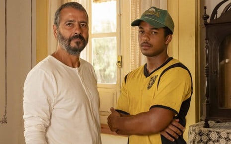 Marcos Palmeira usa blusa branca e posa de frente para Juan Paiva, que está de boné e blusa amarela; eles estão caracterizados como seus personagens em Renascer