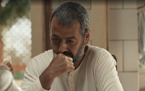 Em cena de Renascer, Marcos Palmeira está com a expressão séria, com a boca apoiada na mão; ele usa blusa branca