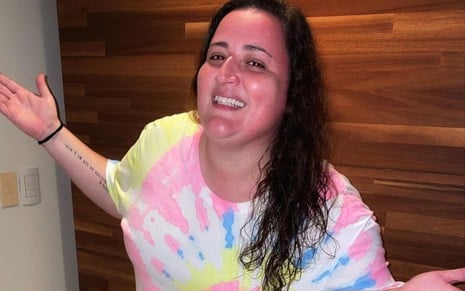 Ju Amaral está com os braços abertos e sorri para foto; ela usa camiseta tie dye