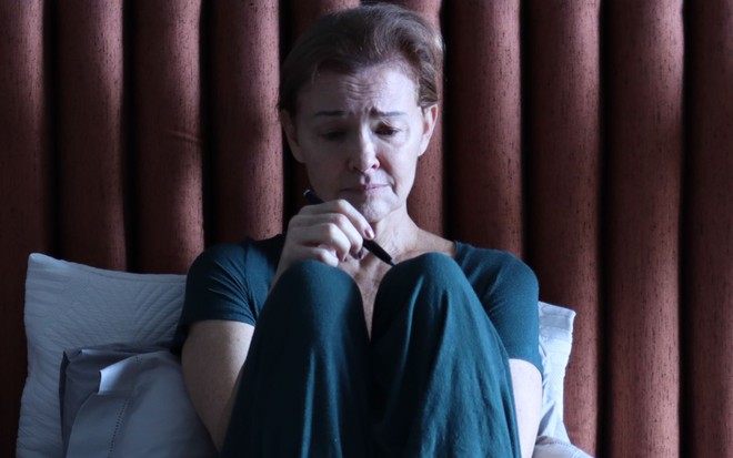 A atriz Julia Lemmertz sentada numa cama, com expressão preocupada
