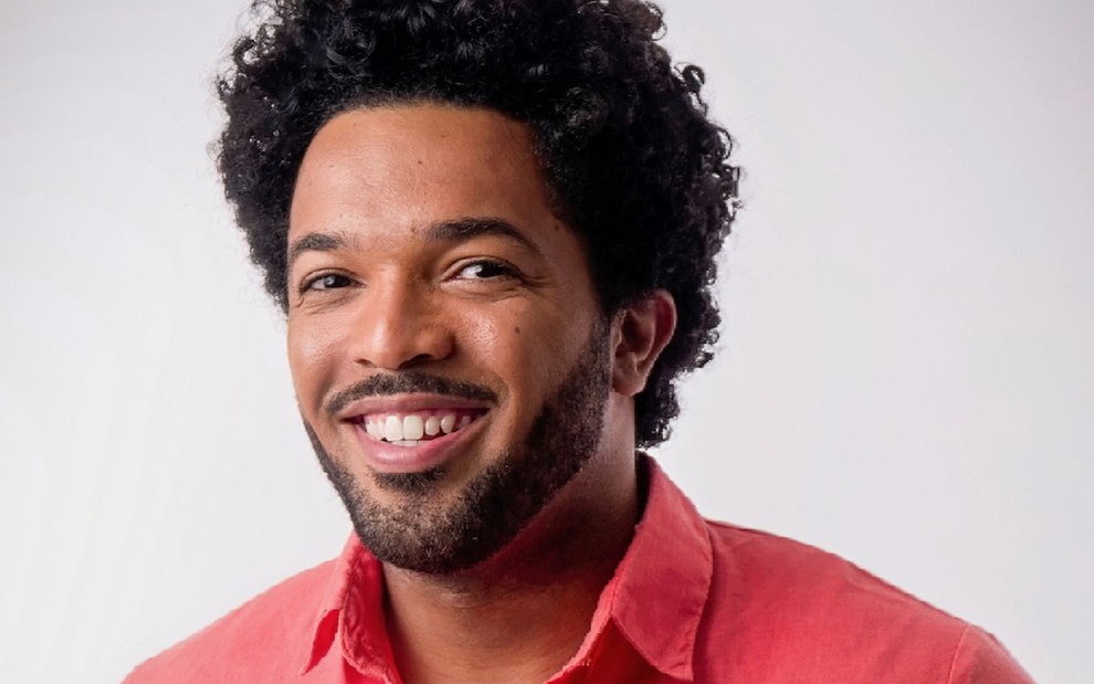 O ator Junior Vieira, com uma camisa vermelha, sorri em frente a uma parede branca