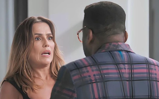 Em cena de Elas por Elas, Deborah Secco está com a expressão de susto olhando para Lázaro Ramos, que está de costas; ele veste blazer xadrez