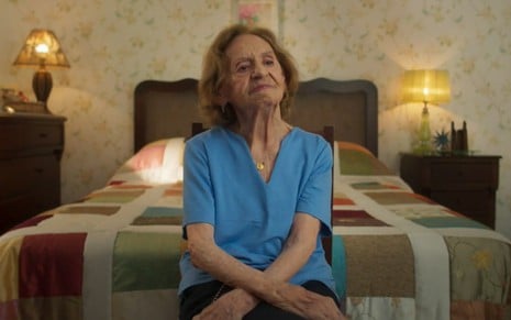 A atriz Laura Cardoso com expressão séria, sentada em frente à cama em seu quarto, no programa Tributo a Laura Cardoso