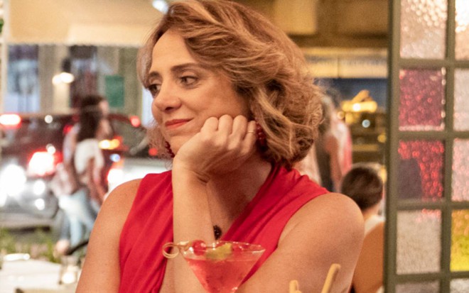 A atriz Leticia Isnard na novela Amor de Mãe, da Globo, com mão no queixo em cenário de bar, séria