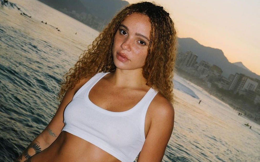 Letícia Vieira tem cabelo cacheado ruivo e está na praia com um top branco