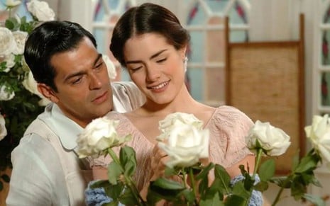 Os atores Eduardo Moscovis e Liliana de Castro ao lado um do outro, ela mexendo em rosas, em cena de Alma Gêmea