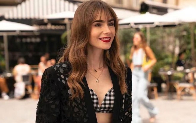 Lily Collins sorri, com um terno preto e um cropped, em cena da série Emily em Paris