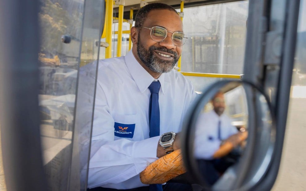 O ator MV Bill sorrindo em cena como Lindomar, dentro de ônibus, em Volta por Cima