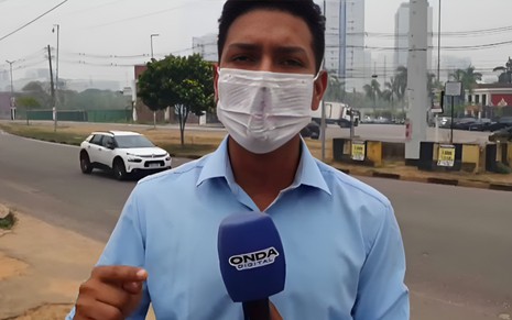 Lucas Conrado está de máscara, em frente a uma rua, e segura o microfone