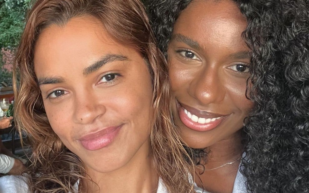 Duas mulheres sorrindo em um close-up, uma com cabelo ondulado castanho claro e a outra com cabelo cacheado preto. Elas estão juntas e parecem felizes