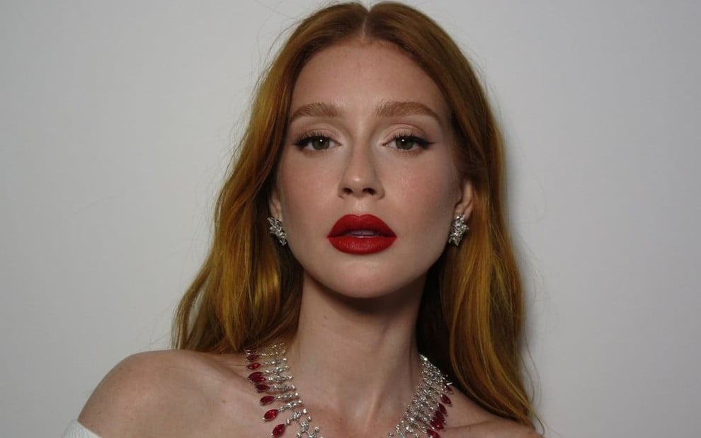 Marina Ruy Barbosa está maquiada e usa um batom vermelho escuro, da mesma cor das pedras de um colar; ela encara a câmera