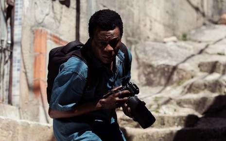 Alexandre Rodrigues corre enquanto segura uma câmera fotográfica em cena da série Cidade de Deus: A Luta Não Para