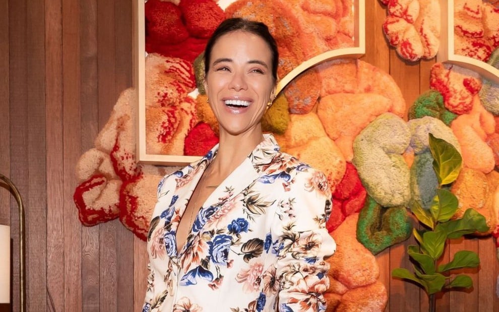 Na imagem, uma mulher sorridente usa um blazer florido, posando em frente a uma parede com texturas e cores vibrantes como laranja, vermelho e verde.