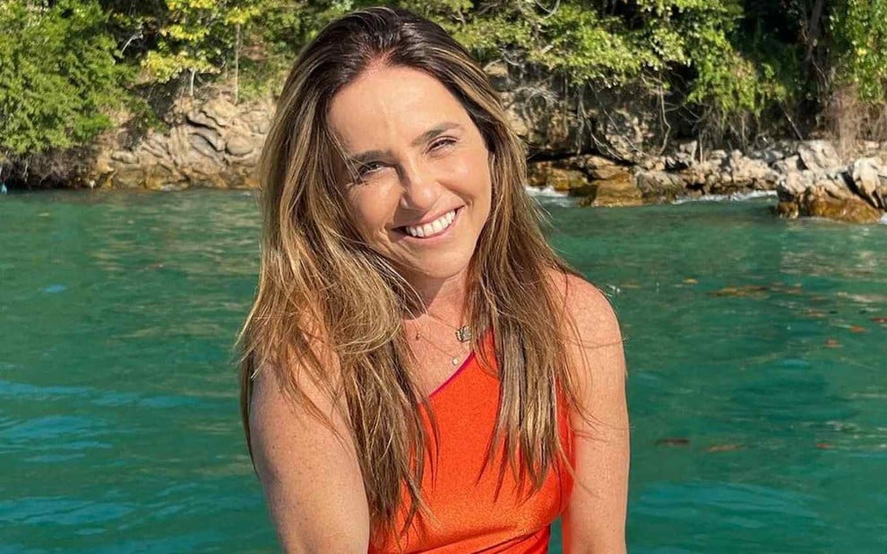 A atriz Monique Curi sorrindo em foto publicada em seu perfil no Instagram, de maiô laranja, em frente ao mar