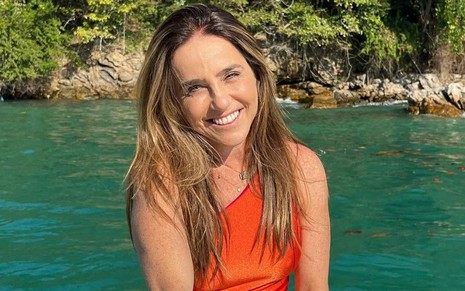 A atriz Monique Curi sorrindo em foto publicada em seu perfil no Instagram, de maiô laranja, em frente ao mar