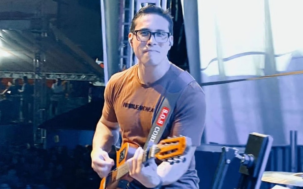 Ayres Sasaki está no palco; ele usa uma camiseta cinza e toca guitarra