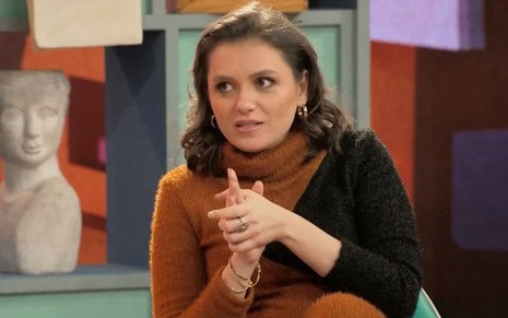 Monica iozzi no Portugal Show; ela está séria e usa uma blusa de tricô marrom e preta