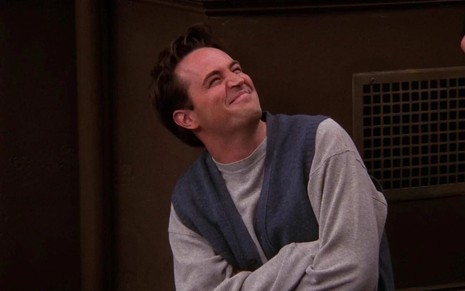 Matthew Perry olha para cima e sorri forçadamente em cena de Friends