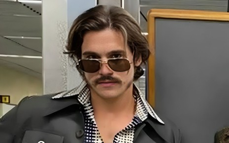 Nicolas Prattes posa com jaquetra preta, blusa quadriculada, usa óculos de sol, bigode e costeleta