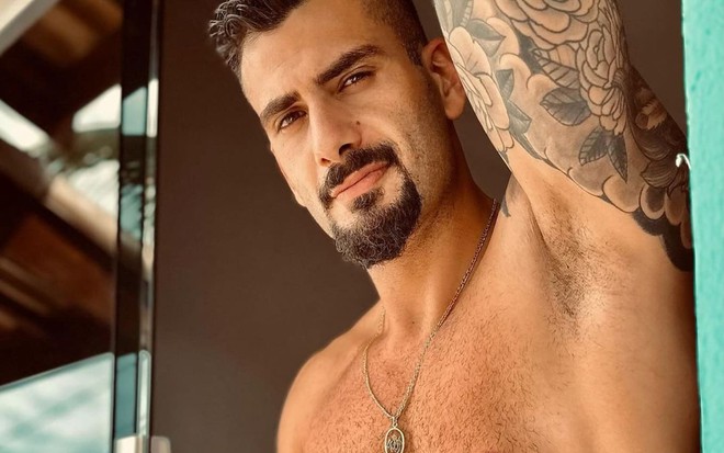 Nizam Hayek está sem camisa e se apoia numa janela enquanto encara a câmera