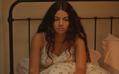 A atriz Larissa Bocchino com expressão séria, sentada na cama de camisola, em cena como Quinota na novela No Rancho Fundo
