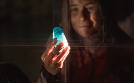 A atriz Andrea Beltrão no plano de fundo, segurando uma pedra azul brilhante em cena de No Rancho Fundo