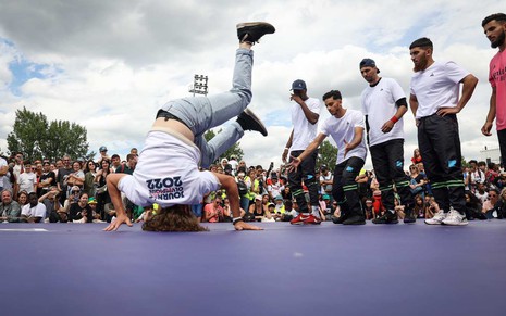 Atletas fazem demonstração de break em rua de Paris; na imagem, um jovem está de ponta-cabeça, enquanto os outros observam