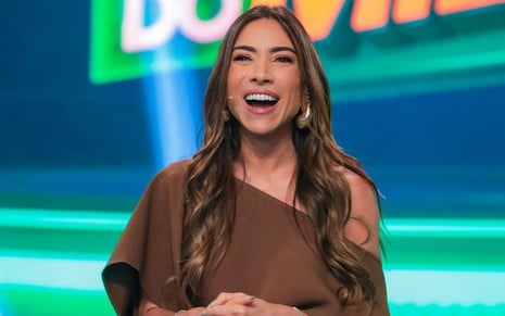 Patricia Abravanel sorridente no quadro Show do Milhão; ela está de cabelos soltos e usa vestido marrom