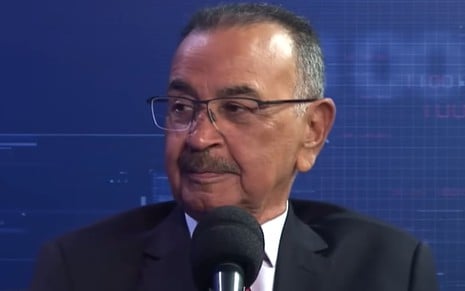 Percival de Souza usa óculos de grau e terno enquanto fala num microfone