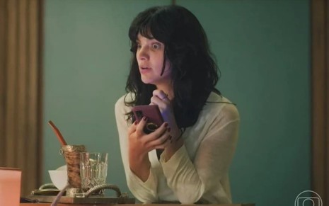 A atriz Debora Ozório segurando celular, com expressão muito assustada em cena de Terra e Paixão