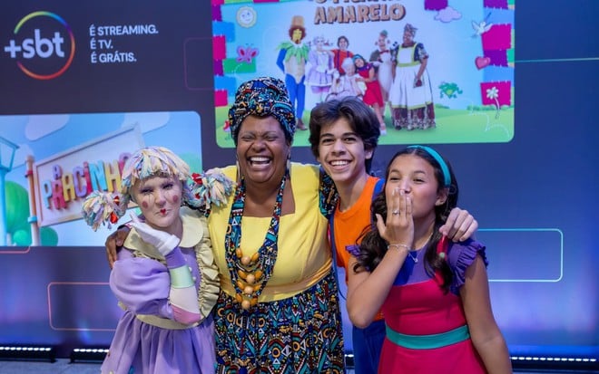 Débora Gomez, Bibba Chuqui, Felipe Lago e Marianna Santos estão abraçados, na frente de um banner do +SBT com fotos de divulgação de Picapau Amarelo e de A Pracinha