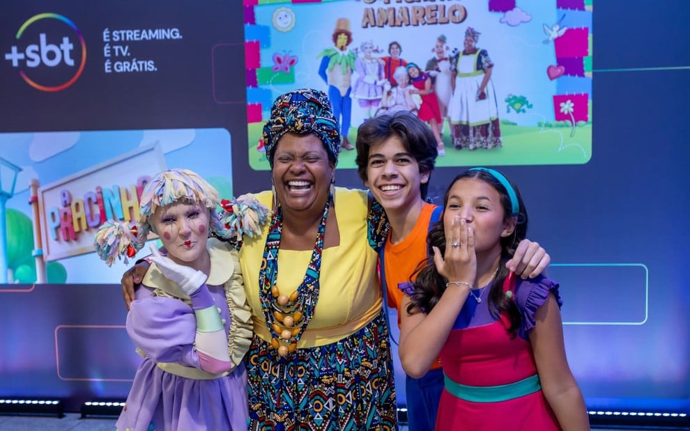 Débora Gomez, Bibba Chuqui, Felipe Lago e Marianna Santos estão abraçados, na frente de um banner do +SBT com fotos de divulgação de Picapau Amarelo e de A Pracinha