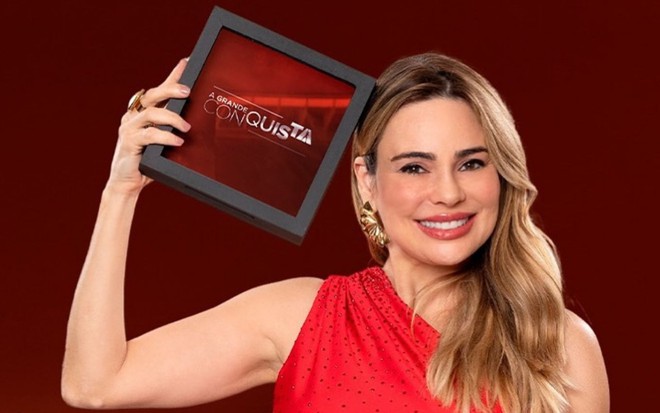 Rachel Sheherazade está com vestido vermelho e segura um quadro com o logo do reality A Grande Conquista