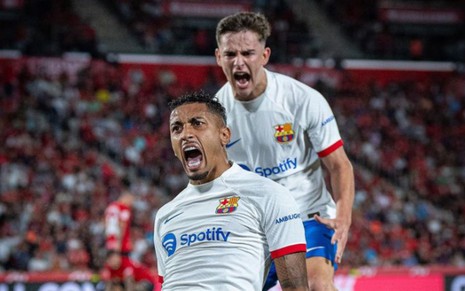 Raphinha (ajoelhado) e Gavi comemoram gol do Barcelona; eles usam uniforme branco e azul do clube