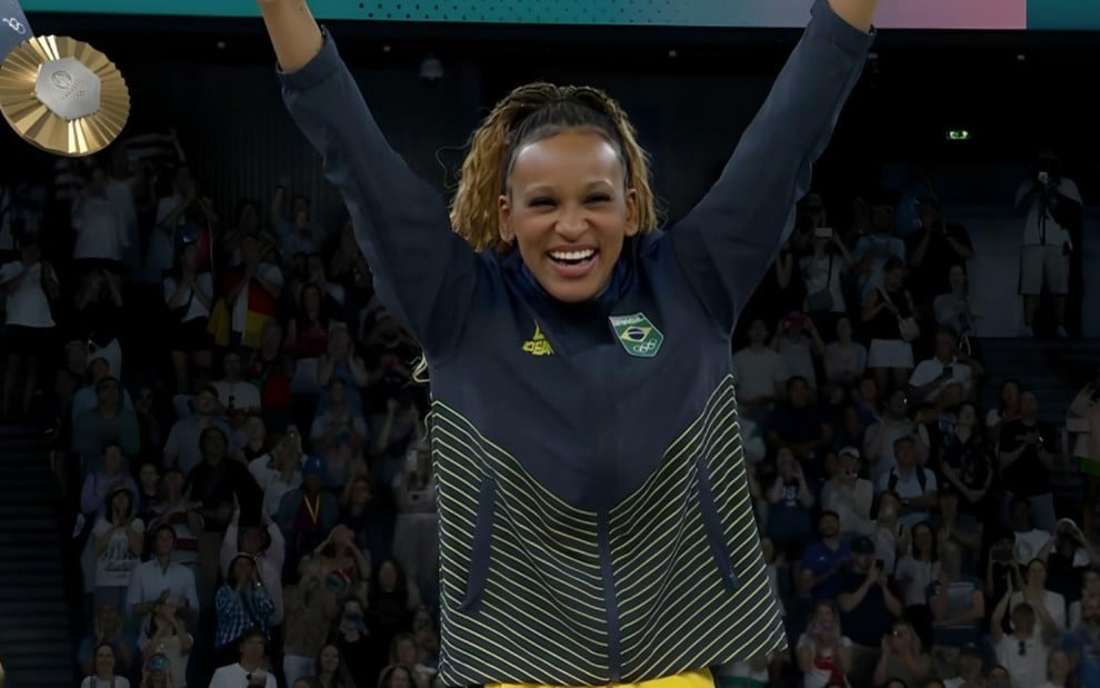 Rebeca Andrade com os braços para cima, sorrindo, no momento em que subiu ao pódio para receber medalha de ouro na Olimpíada de Paris, na segunda (5)