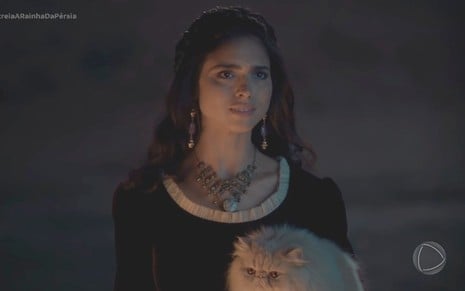 Nathalia Florentino tem expressão confusa e segura um gato branco em cena de A Rainha da Pérsia