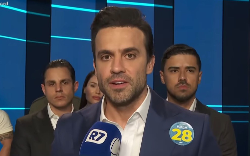 A imagem mostra um homem de terno azul e camisa branca segurando um microfone azul com o logo "R7". Ele usa um broche com o número 28.