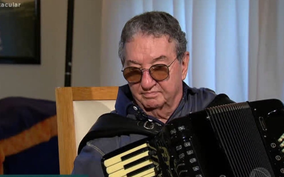 O músico Caçulinha de óculos escuros com um acordeão no colo em entrevista à Record