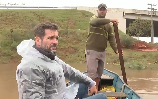 Romeu Piccoli tem expressão abalada enquanto navega por rio alagado; ele usa uma capa de chuva