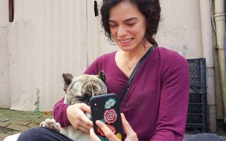 Uma mulher de cabelo escuro e cacheado, usando blusa roxa, está sentada sorrindo enquanto segura um cachorro e tira uma selfie com o celular.